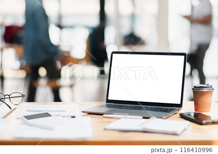 A laptop with a blank screen, coffee cup, and office supplies on a wooden desk in a modern open office setting with blurred background. 116410996