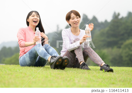 運動後に芝生の上で水分補給する二人の女性 運動後に芝生の上で水分補給する二人の女性 116411413