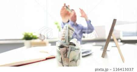 Rich man throwing US banknotes in big jar full of money at workplace 116413037
