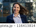 Portrait of an attractive young adult woman in a blue suit standing against the background of an office building, smiling and looking at the camera close-up 116413814