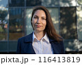 Portrait of an attractive strong confident independent woman in a blue suit standing against the background of a business building and looking at the camera 116413819