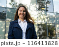 Portrait of a happy caucasian brunette woman in a blue suit stands against the background of a business building and smiles 116413828