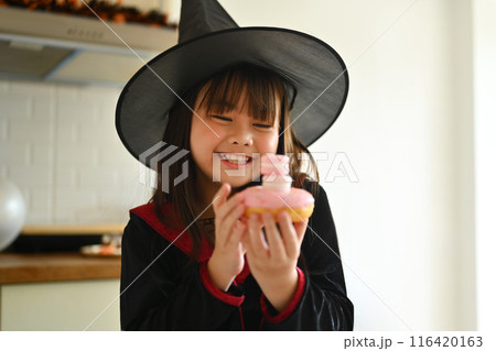 Portrait of cute little girl in witch costumes holding Halloween desserts standing in decorated room Portrait of cute little girl in witch costumes holding Halloween desserts standing in decorated room 116420163