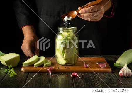 A cook spoons salt into a jar of chopped vegetable marrow before canning it with dill and garlic. Concept of pickling delicious zucchini on the kitchen table A cook spoons salt into a jar of chopped vegetable marrow before canning it with dill and garlic. Concept of pickling delicious zucchini on the kitchen table 116421275