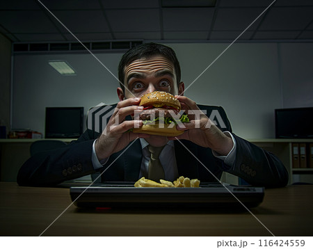 photography of a man in an office eating, there's a laptop on the desk in front of him	 116424559