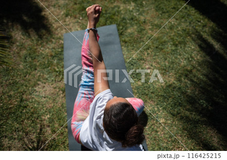 Woman practicing yoga outdoors on a mat, wearing colorful leggings and white top, performing a seated twist posture in a sunny garden Woman practicing yoga outdoors on a mat, wearing colorful leggings and white top, performing a seated twist posture in a sunny garden 116425215