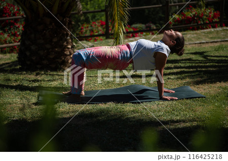 Woman practicing yoga outdoors on a sunny day performing reverse plank pose on a yoga mat in a serene garden setting Woman practicing yoga outdoors on a sunny day performing reverse plank pose on a yoga mat in a serene garden setting 116425218