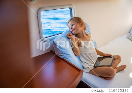 Little girl sits with tablet in the cabin of a yacht and looks out the window during a sea crossing. Yacht life concept 116425382