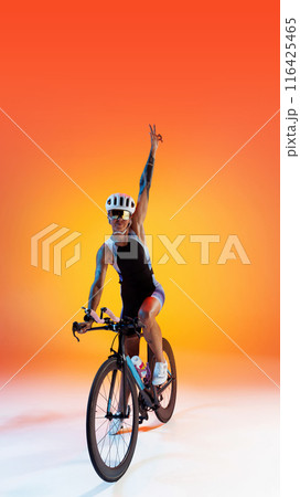 Champion, winner. Woman in sportswear and helmet sitting on bike and raising hands upward in victory pose against gradient background in neon light 116425465
