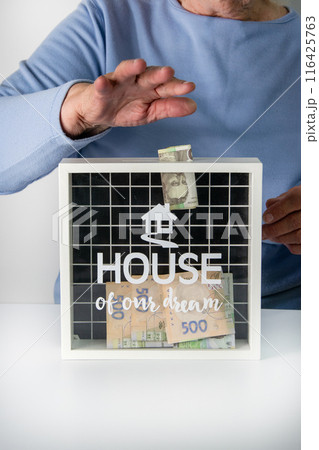 An elderly person in a blue shirt is saving money in a white jar labeled House of our dream 116425763