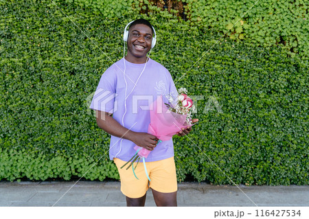 Happy African man with headphones wearing a purple shirt is holding a bouquet of flowers. He is wearing headphones and he is listening to music 116427534