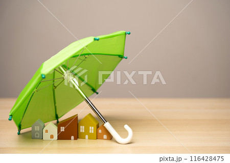 Property and home insurance concept. Wooden houses stand under a green umbrella. Financial security. Repairs or rebuilding in the event of a covered loss. Property and home insurance concept. Wooden houses stand under a green umbrella. Financial security. Repairs or rebuilding in the event of a covered loss. 116428475