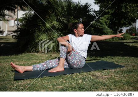 Woman practicing yoga outdoors on a sunny day, performing seated twist pose on a yoga mat surrounded by lush greenery 116430897