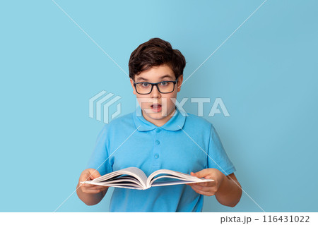 A surprised schoolboy in glasses, holding an open book in his hands, look at camera 116431022