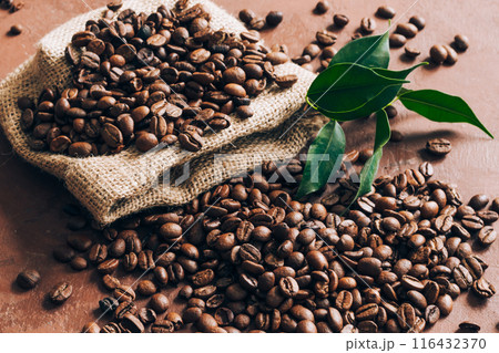 Burlap bag scattering coffee beans with green leaves on table. Top view, flat lay Burlap bag scattering coffee beans with green leaves on table. Top view, flat lay 116432370