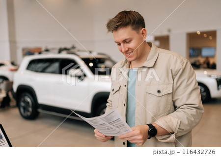 Happy young male customer holding papers, checking vehicle characteristics during choosing car in modern dealership center, smiling looking down. Concept of choosing and buying new auto at showroom Happy young male customer holding papers, checking vehicle characteristics during choosing car in modern dealership center, smiling looking down. Concept of choosing and buying new auto at showroom 116432726