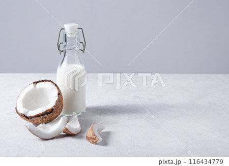 Glass vegan milk bottle on white marble table with half coconuts. Front view and copy space 116434779