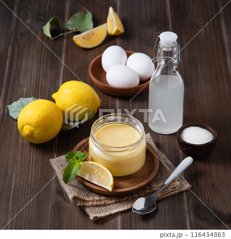 Delicious lemon curd in a jar on a dark wooden background with fresh citrus fruits and ingredients.  116434863