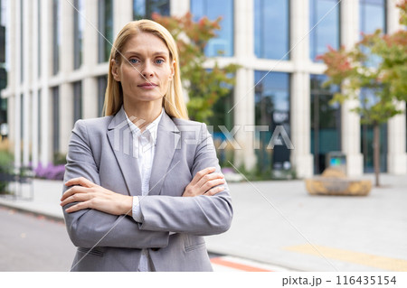 Confident businesswoman in gray suit standing outside modern office building with arms crossed. Professional and determined expression. Successful corporate woman in outdoor urban setting. Confident businesswoman in gray suit standing outside modern office building with arms crossed. Professional and determined expression. Successful corporate woman in outdoor urban setting. 116435154