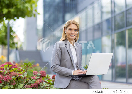 Confident businesswoman working on laptop outdoors in modern urban environment. Professional in formal attire seated outside office building surrounded by greenery, representing successful career Confident businesswoman working on laptop outdoors in modern urban environment. Professional in formal attire seated outside office building surrounded by greenery, representing successful career 116435163