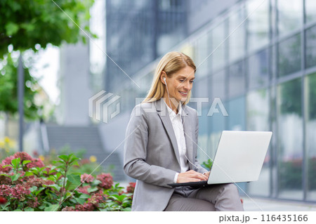 Businesswoman using laptop outdoors with earphones in urban setting. Professional attire in business environment. Concept of remote work, technology, and communication. Businesswoman using laptop outdoors with earphones in urban setting. Professional attire in business environment. Concept of remote work, technology, and communication. 116435166