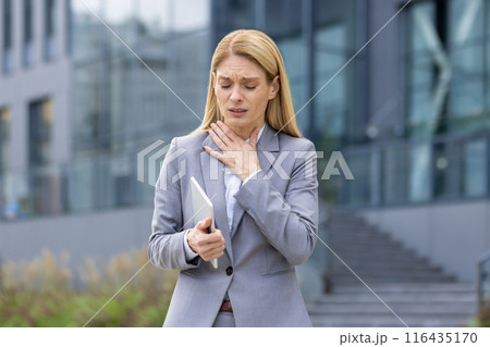 Businesswoman in gray suit holding tablet outside modern office building, experiencing throat discomfort. Scene portrays stress, pain, and health issues in a business environment. Businesswoman in gray suit holding tablet outside modern office building, experiencing throat discomfort. Scene portrays stress, pain, and health issues in a business environment. 116435170