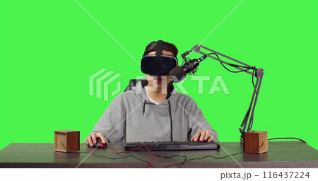 Video blogger recording live gameplay at desk, playing online tournament with virtual reality interactive headset. Asian man streaming videogames vlog, content creator on greenscreen. 116437224