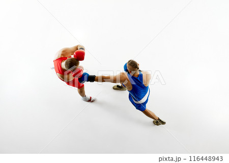 Aerial view of two men boxing, showcasing blue fighter's punch aimed at fighter in red uniform against white studio background. 116448943