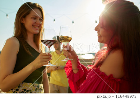 Photo of lively toast among friends, two beautiful women. Their faces lit up with smiles and drinks sparkling in sun. 116448975