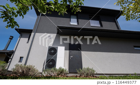 Positioned by the wall of a modern cottage, the outdoor unit of a heat pump is visible. 116449377