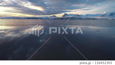 Low angle flight over polar icy ocean in Antarctica. Snow-covered mountains range under colorful sunset clouds sky. Tranquil scene icy landscape, wild nature background. South Pole travel, exploration 116452305