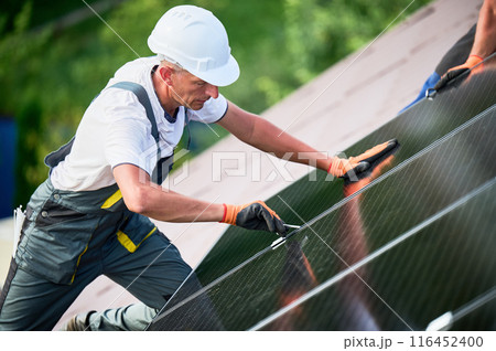 Worker building photovoltaic solar panel system on rooftop of house. Man engineer in helmets and gloves installing solar module with help of hex key outdoors. Alternative energy generation concept. Worker building photovoltaic solar panel system on rooftop of house. Man engineer in helmets and gloves installing solar module with help of hex key outdoors. Alternative energy generation concept. 116452400