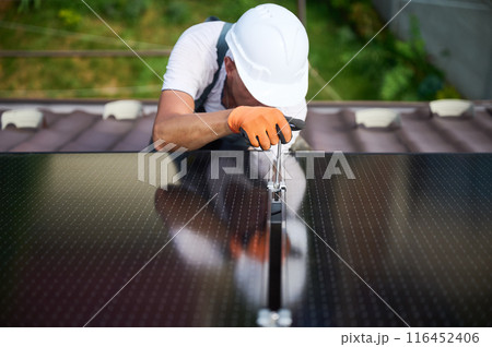 Worker building photovoltaic solar panel system on rooftop of house. Close up of man engineer in helmets and gloves installing solar module with help of hex key outdoors. Alternative, renewable energy 116452406