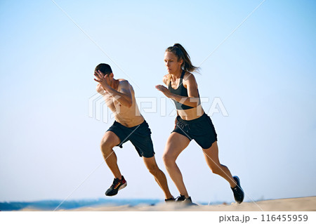 Dynamic and hard training outdoor session. Sportive young man and woman training on sandy beach, running under clear sky. 116455959