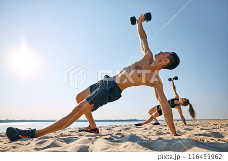 Man and woman performing side plank with dumbbell lifts on sandy beach, under clear blue sky. Full body outdoor workout 116455962