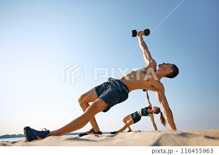 Man and woman performing side plank with dumbbell lifts on sandy beach, under clear blue sky. Full body outdoor workout 116455963