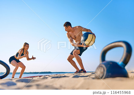 Man and woman exercising on sandy beach with medicine ball, with under a clear blue sky. Intensity functional training Man and woman exercising on sandy beach with medicine ball, with under a clear blue sky. Intensity functional training 116455968