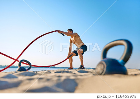 Muscular shirtless man in black shorts training with battle ropes on sandy beach under blue clear sky, with kettle bell lying in sand. 116455978