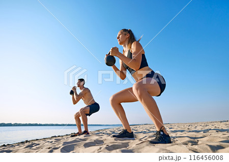 Sportive young man and woman with sportive body, in black sportswear doing squats with kettlebell, intensive outdoor training on beach by the river. Sportive young man and woman with sportive body, in black sportswear doing squats with kettlebell, intensive outdoor training on beach by the river. 116456008