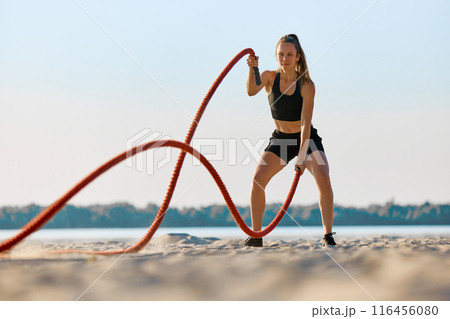 Woman in black sportswear exercising with red battle ropes on a sandy beach, with water and trees in background under clear sky. Woman in black sportswear exercising with red battle ropes on a sandy beach, with water and trees in background under clear sky. 116456080
