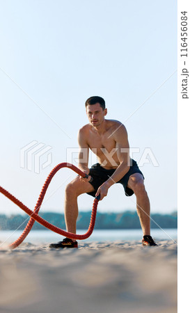 Athlete performing battle rope exercises on sandy beach, shirtless muscular man in n black shorts doing intensity functional training outdoors Athlete performing battle rope exercises on sandy beach, shirtless muscular man in n black shorts doing intensity functional training outdoors 116456084