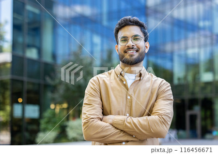Young professional man with glasses standing confidently outside modern glass office building, smiling and crossing his arms. Business and success concept in urban environment. Young professional man with glasses standing confidently outside modern glass office building, smiling and crossing his arms. Business and success concept in urban environment. 116456671