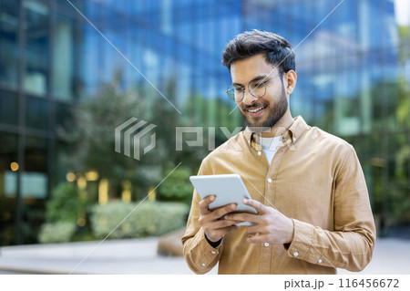 Young man smiling while using tablet outside modern office building. Casual attire, urban setting, and contemporary design. Represents technology, business, and communication. Happy expression 116456672