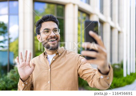 Happy man making video call with smartphone outside modern office building, waving and smiling. Man wearing glasses and casual shirt, enjoying outdoor work environment and connection. Happy man making video call with smartphone outside modern office building, waving and smiling. Man wearing glasses and casual shirt, enjoying outdoor work environment and connection. 116456691