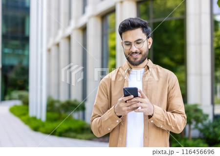 Smiling young man wearing glasses using smartphone outside modern office building. Casually dressed man engaging with social media or business apps phone, bright environment representing technology Smiling young man wearing glasses using smartphone outside modern office building. Casually dressed man engaging with social media or business apps phone, bright environment representing technology 116456696