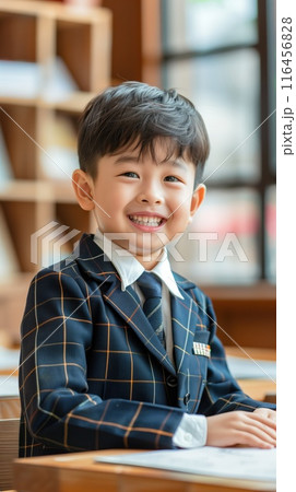 Seven year old Asian boy wears blue orange plaid suit with white collar and black tie. Smiles at camera in classroom with bookshelf full of books suggesting knowledge learning. Back to school theme. 116456828