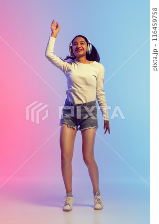 Full-length image of young girl in shorts and t-shirt listening to music in headphones and dancing with joy against gradient pink blue background in neon light 116459758