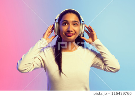 Beautiful young girl in white shirt, listening to music in headphones against gradient pink blue background in neon light 116459760