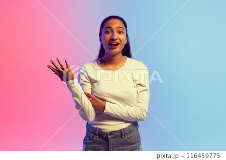 Young woman in white shirt with long black hair, standing against pink and blue gradient background in neon light, with surprised expression. Shock and astonishment. 116459775