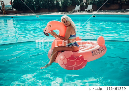 A woman is sitting on a pink inflatable flamingo in a pool. The flamingo is pink and has a blue stripe. The woman is smiling and she is enjoying herself. Concept of fun and relaxation. 116461259